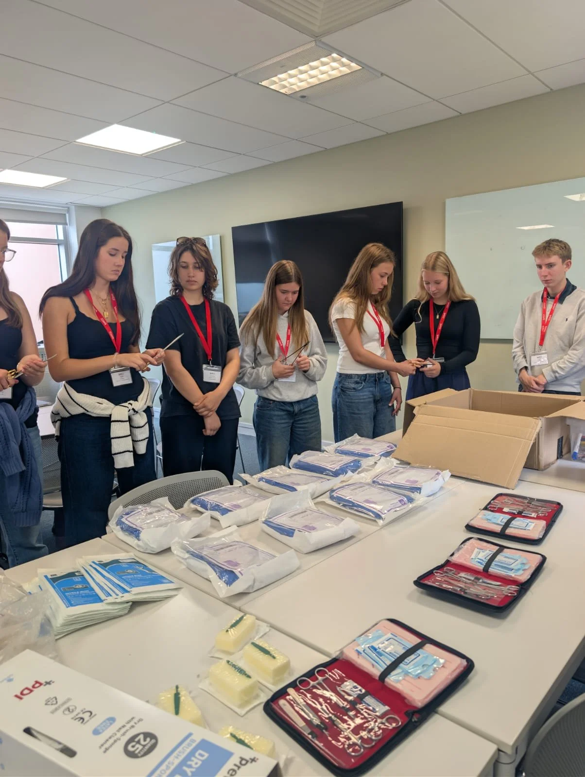 Group of young people standing around a table with dental supplies, including gloves and dental tools, in a classroom or training room.