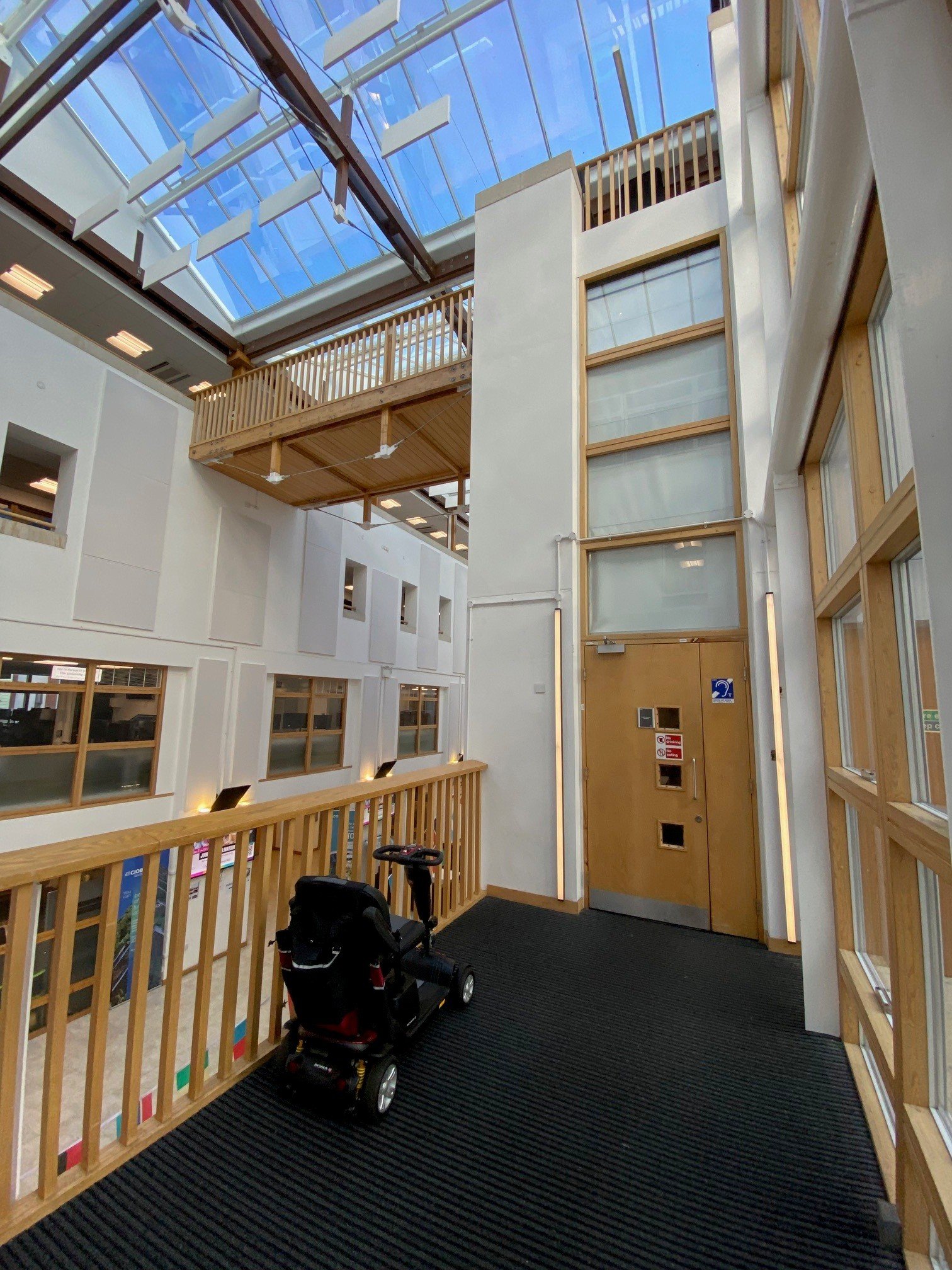 Interior of a modern building in the university of Portsmouth with a glass ceiling, wooden railings, and white walls. A black mobility vehicle is parked near the railing.