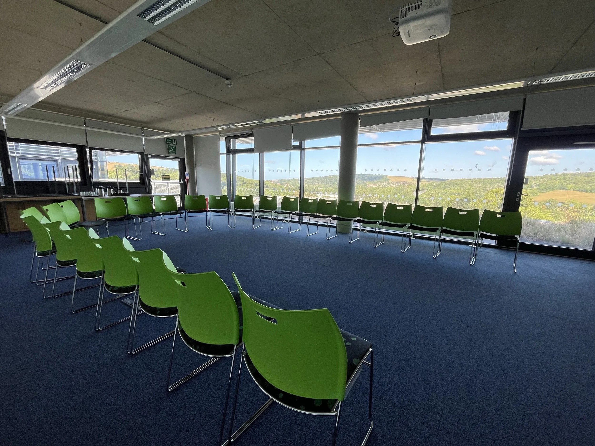 University of Brighton conference room with green chairs arranged in a U-shape in front of large windows overlooking a landscape with hills and blue sky.
