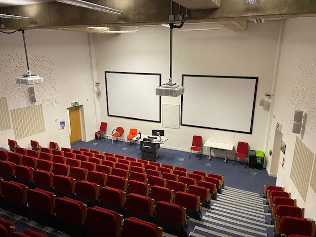 An University of Brighton university lecture hall with red seats, two large projection screens, and a central lectern with computers.