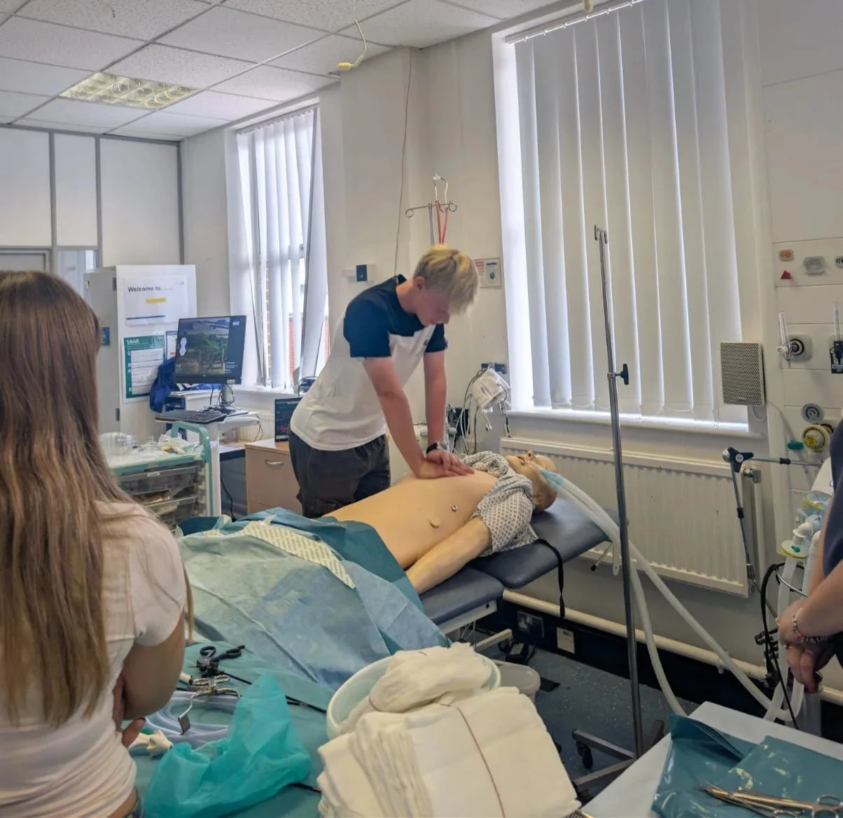 A medical simulation in a hospital room with a mannequin lying on a bed, nurses or students observing, and a work experience pupil practicing chest compressions on the mannequin.