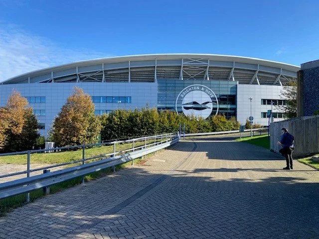 Brighton & Hove Albion football stadium with a large sign featuring a seagull logo, surrounded by trees and a paved walkway.