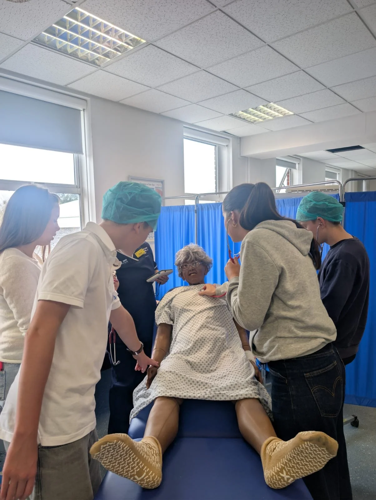 A group of medical students and healthcare professionals examining a mannequin patient lying on a hospital bed in a clinical setting, with blue curtains and windows in the background.