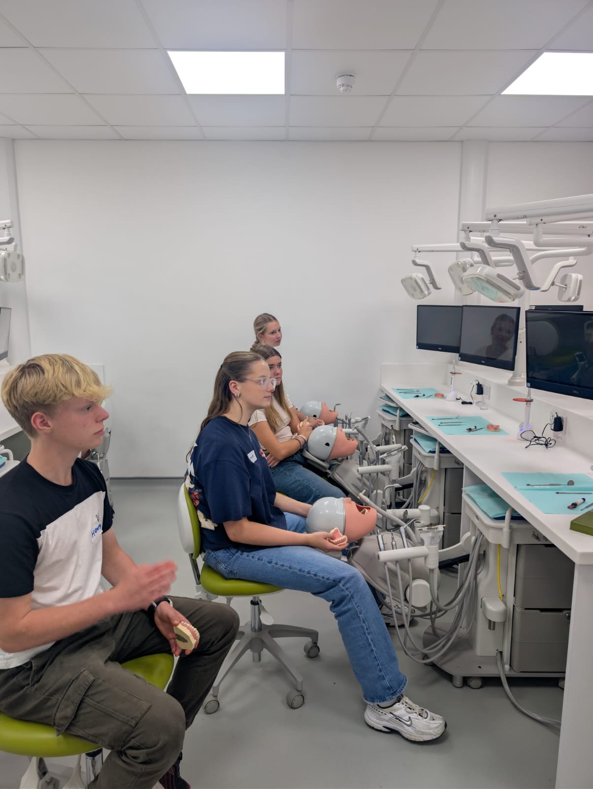 A dental training simulation with four students practicing dental procedures on mannequins in a clinical room with monitors and dental equipment.