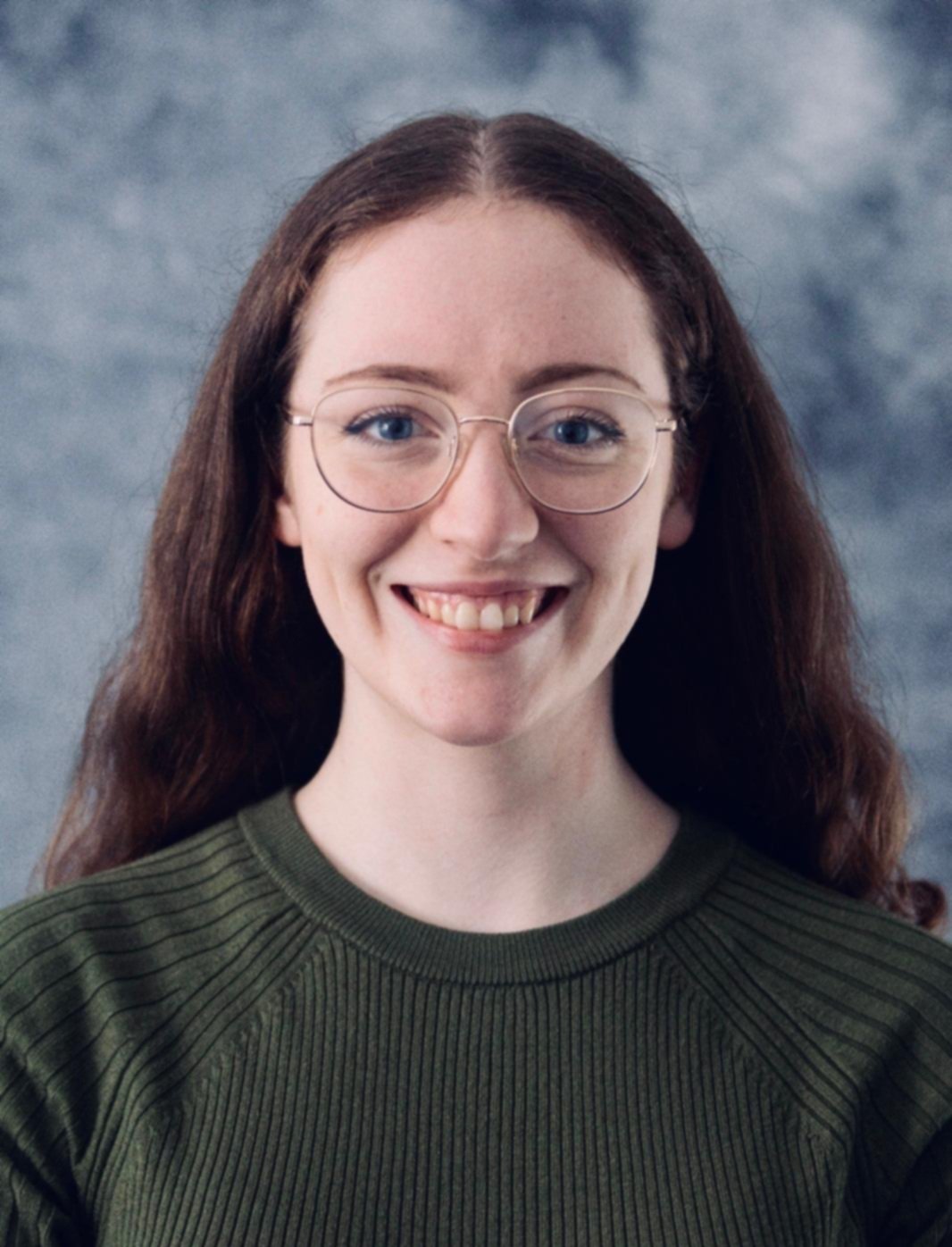 A young woman with long brown hair, glasses, and a big smile, wearing a dark green ribbed top, standing in front of a cloudy blue-gray background.