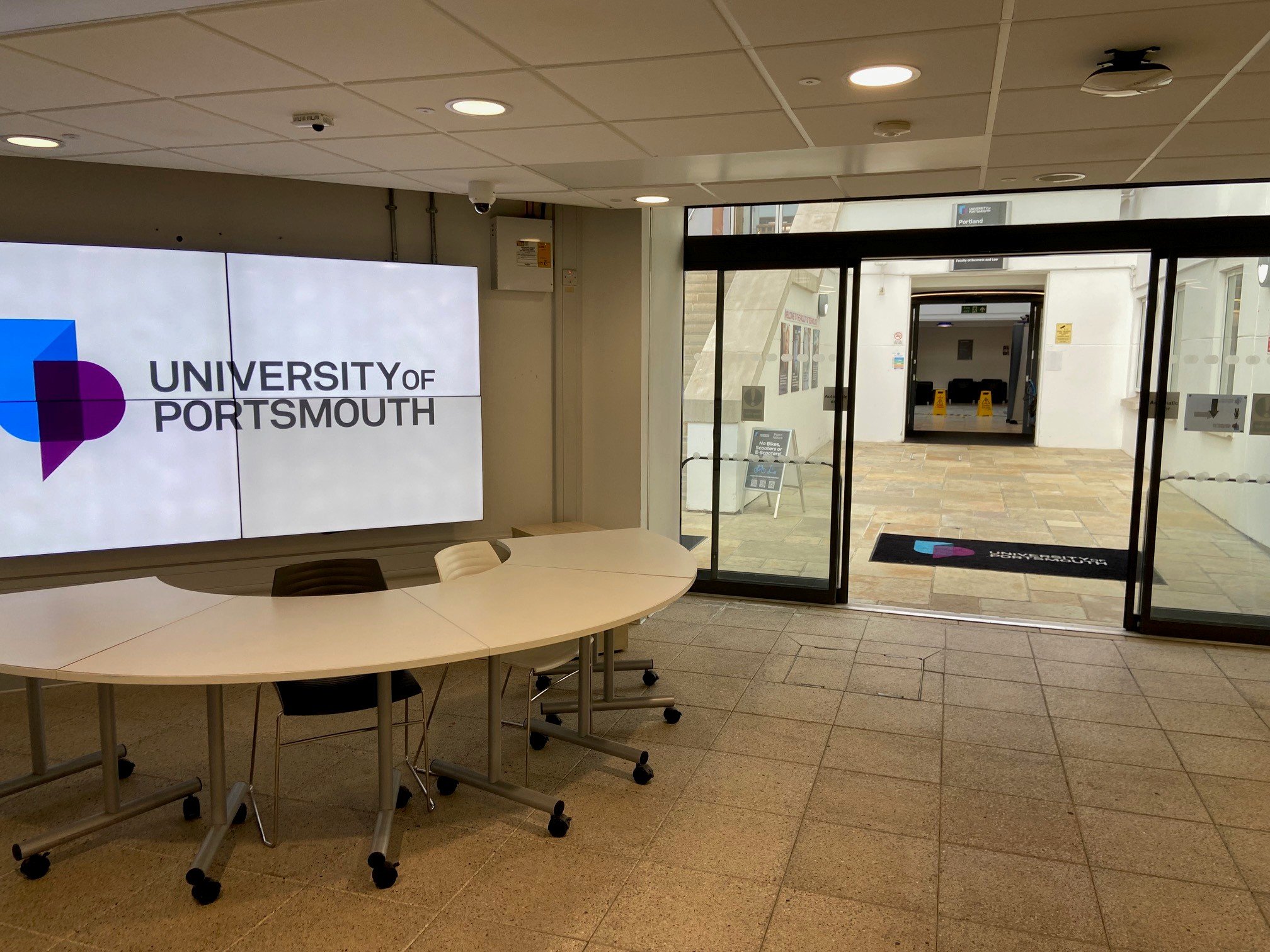 Interior of University of Portsmouth reception area with a large digital screen displaying the university logo and an entrance door leading outside.