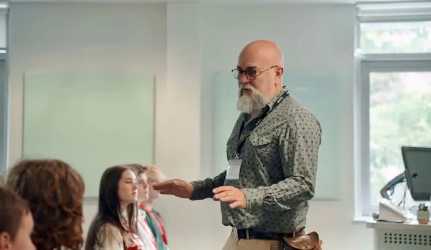 A middle-aged man with a beard and glasses teaches a classroom of young students, gesturing with his hands, in a brightly lit room with large windows.
