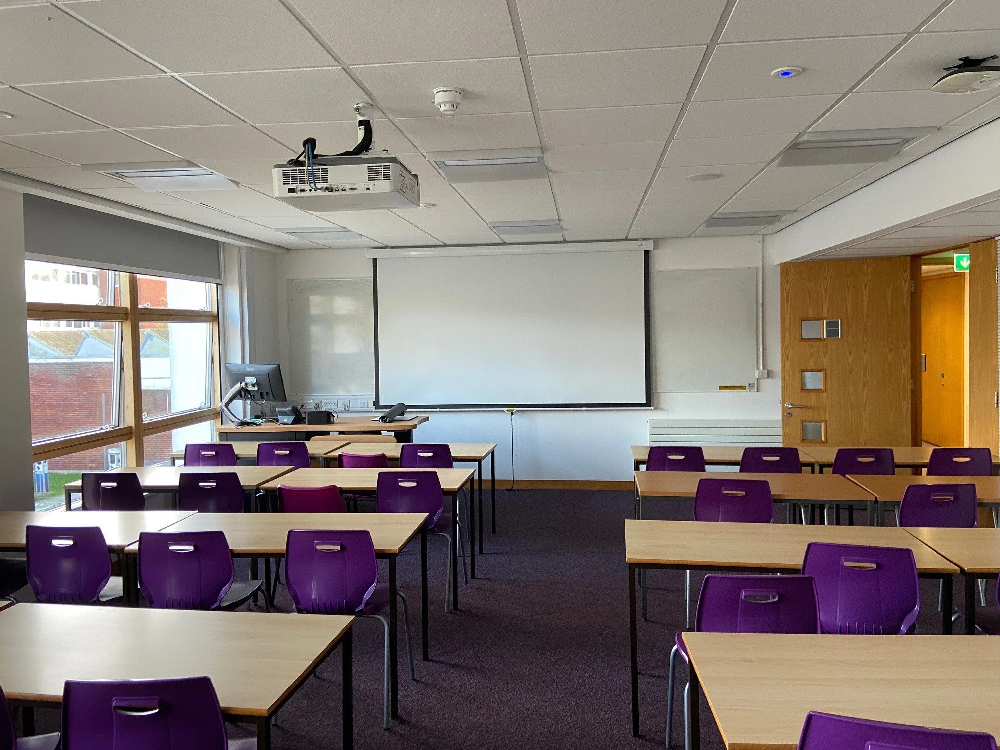 Empty classroom with purple chairs and wooden desks, large white projection screen, windows on the left, and a wooden door on the right.