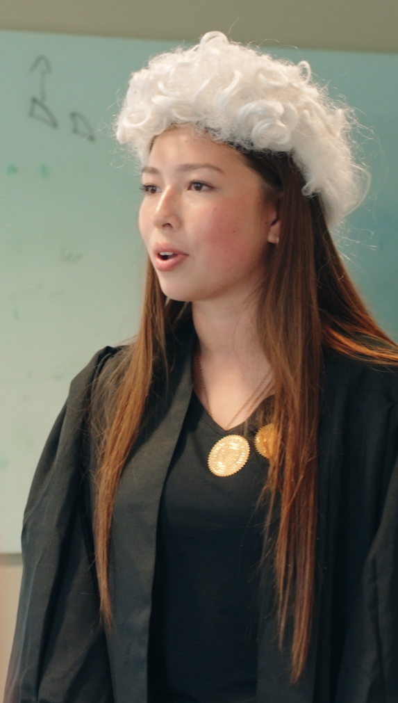 Pupil in a Law work experience mock trial, wearing a white traditional curly wig and a black gown with gold pendant, speaking in front of a whiteboard.
