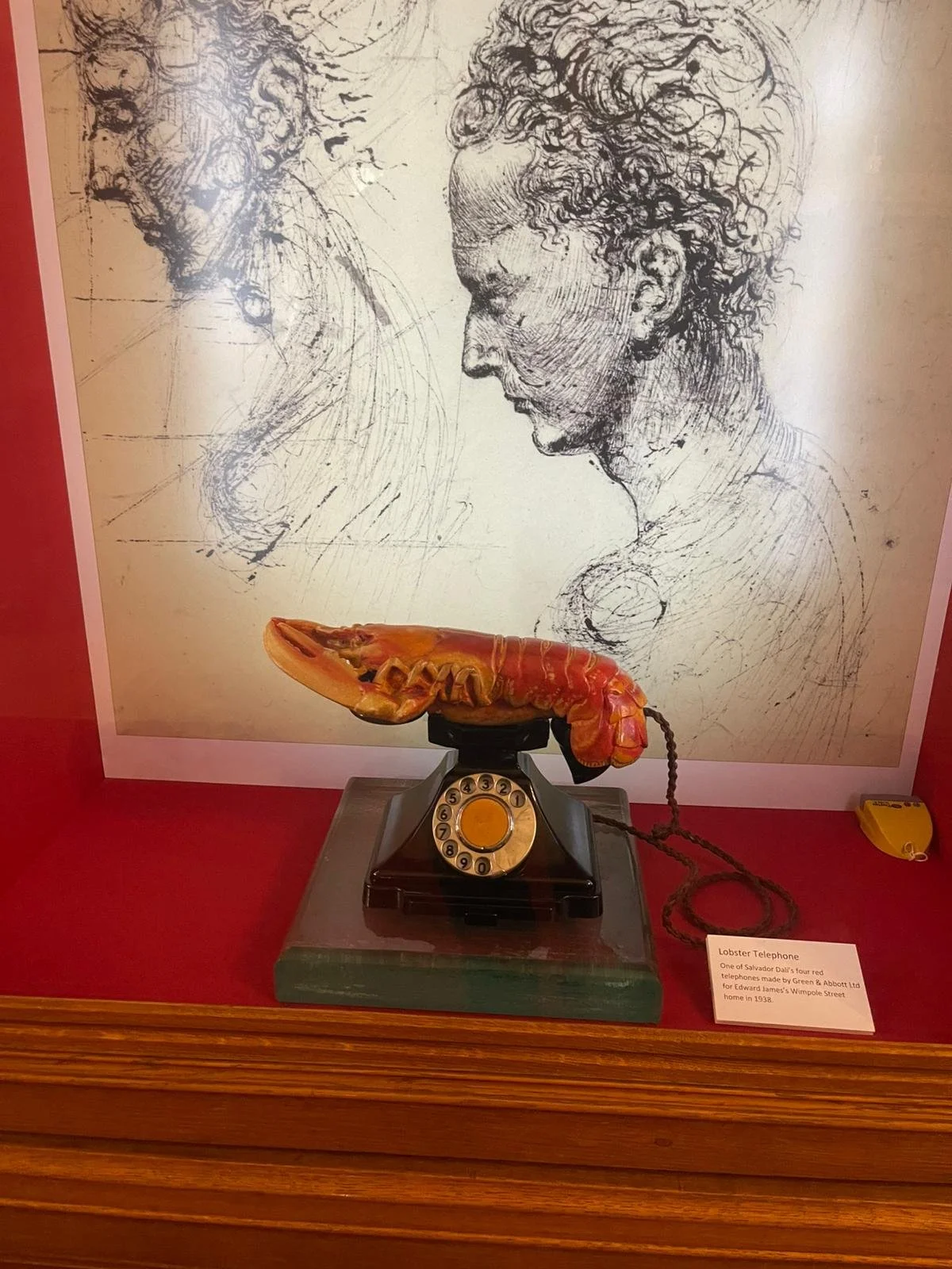Vintage lobster telephone with a red lobster-shaped receiver and cord, displayed on a table with a red background and a sketch drawing of two profiles of a man and a woman in the background.