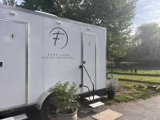 A white mobile food trailer with a logo and the text 'Foxy Looks' on the side, parked outdoors near trees and a fence, with steps leading up to the door.