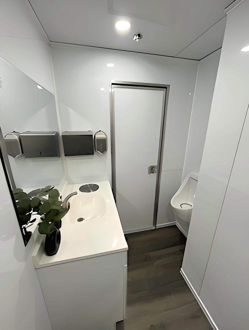 Small modern bathroom with white walls, a sink with a black bottle and green plant, a mirror, two paper towel dispensers, a door, and a urinal.