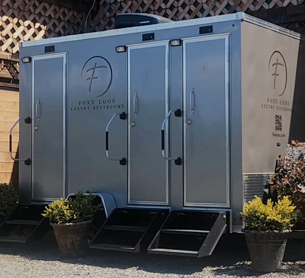 Luxury portable restroom trailer with two doors, steps, and potted plants on either side, labeled 'Foxy Loos'.