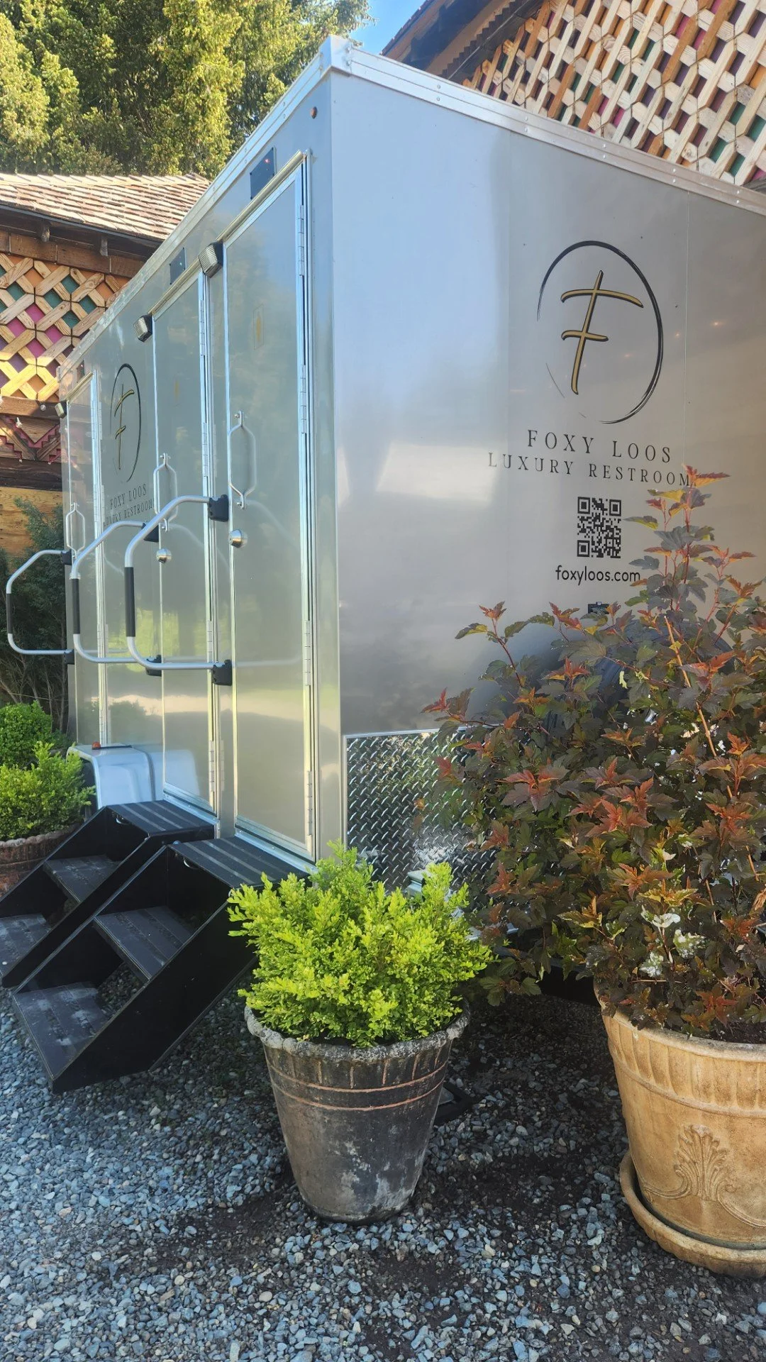 A mobile luxury restroom trailer with stairs, surrounded by potted plants, with trees and a wooden building in the background.