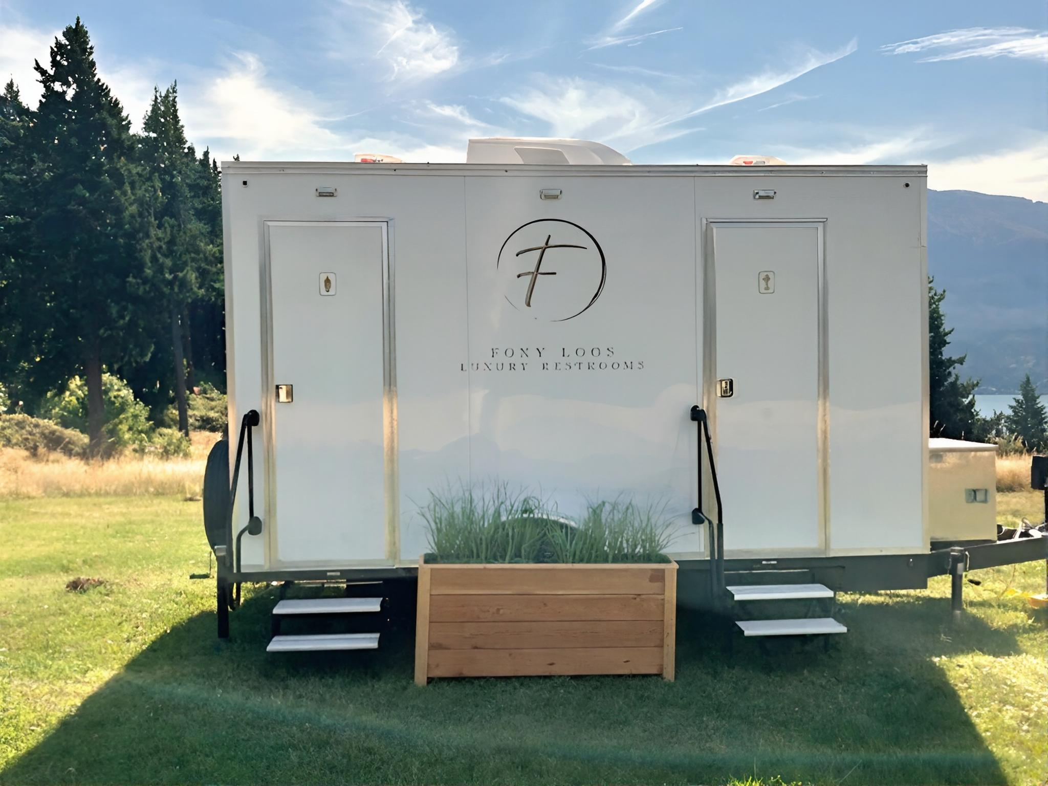 White luxury restroom trailer with black steps leading to two doors, one on each side, set outdoors on grass with trees and mountains in the background, and a planter box with grass in front.