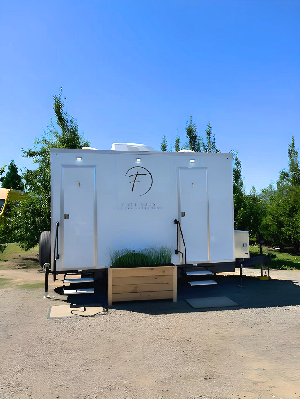 A white food trailer with logo and text, situated outdoors on a sunny day, with trees in the background.
