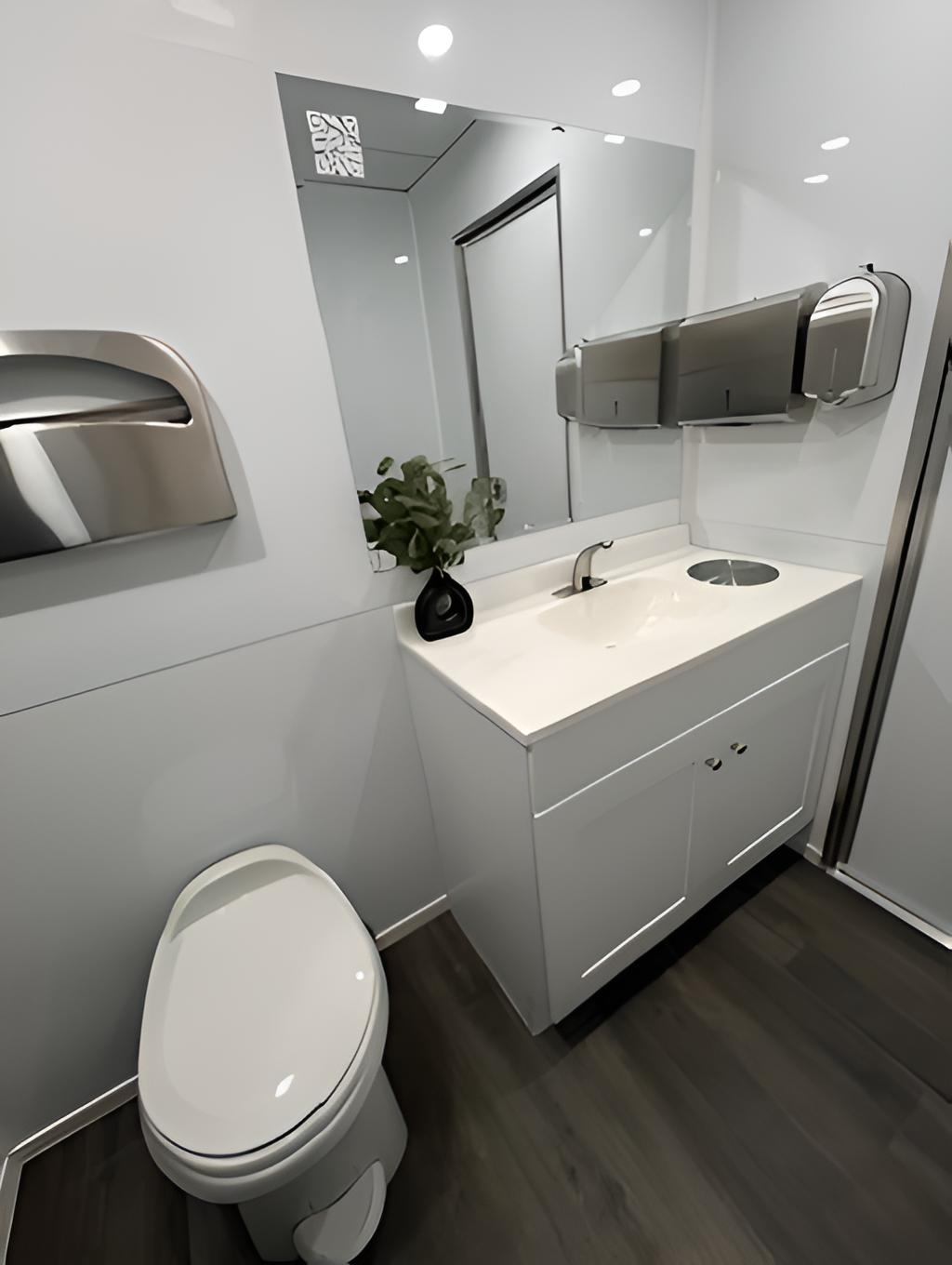 A modern bathroom with a white vanity, sink, and cabinet, a large mirror, a wall-mounted soap dispenser, a toilet, and a potted plant, with a gray wood floor and metallic fixtures.