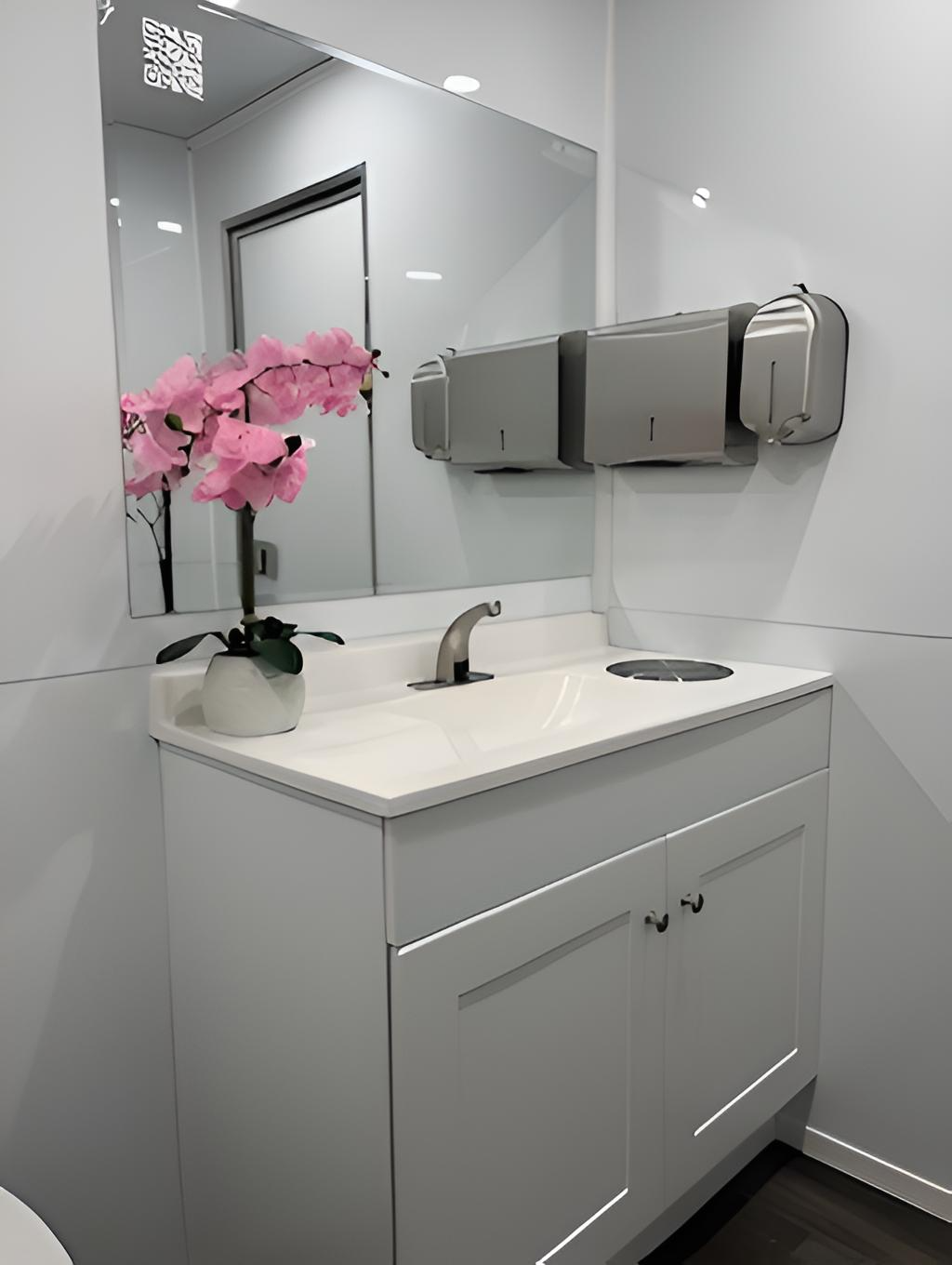 A white bathroom vanity with a sink and faucet, a mirror, a gallon-sized white vase with pink artificial flowers, and a wall-mounted stainless steel paper towel dispenser, with a reflection of a door and ceiling light in the mirror.