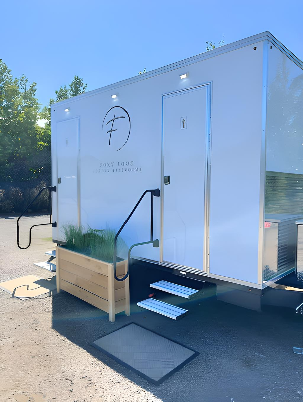 A white portable restroom trailer with a logo, steps, a planter box, and outdoor lighting, set against a backdrop of trees and a clear blue sky.