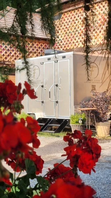 Portable outdoor toilet in a backyard, partially obscured by red flowers and green foliage.