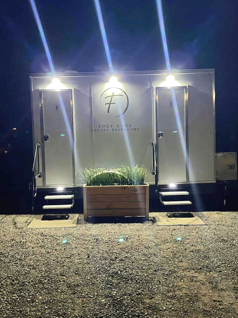 A portable restroom building with two doors, steps, and patio lighting, with a logo and the text "FOXY LOGO BEAUTY BAR" on the front.