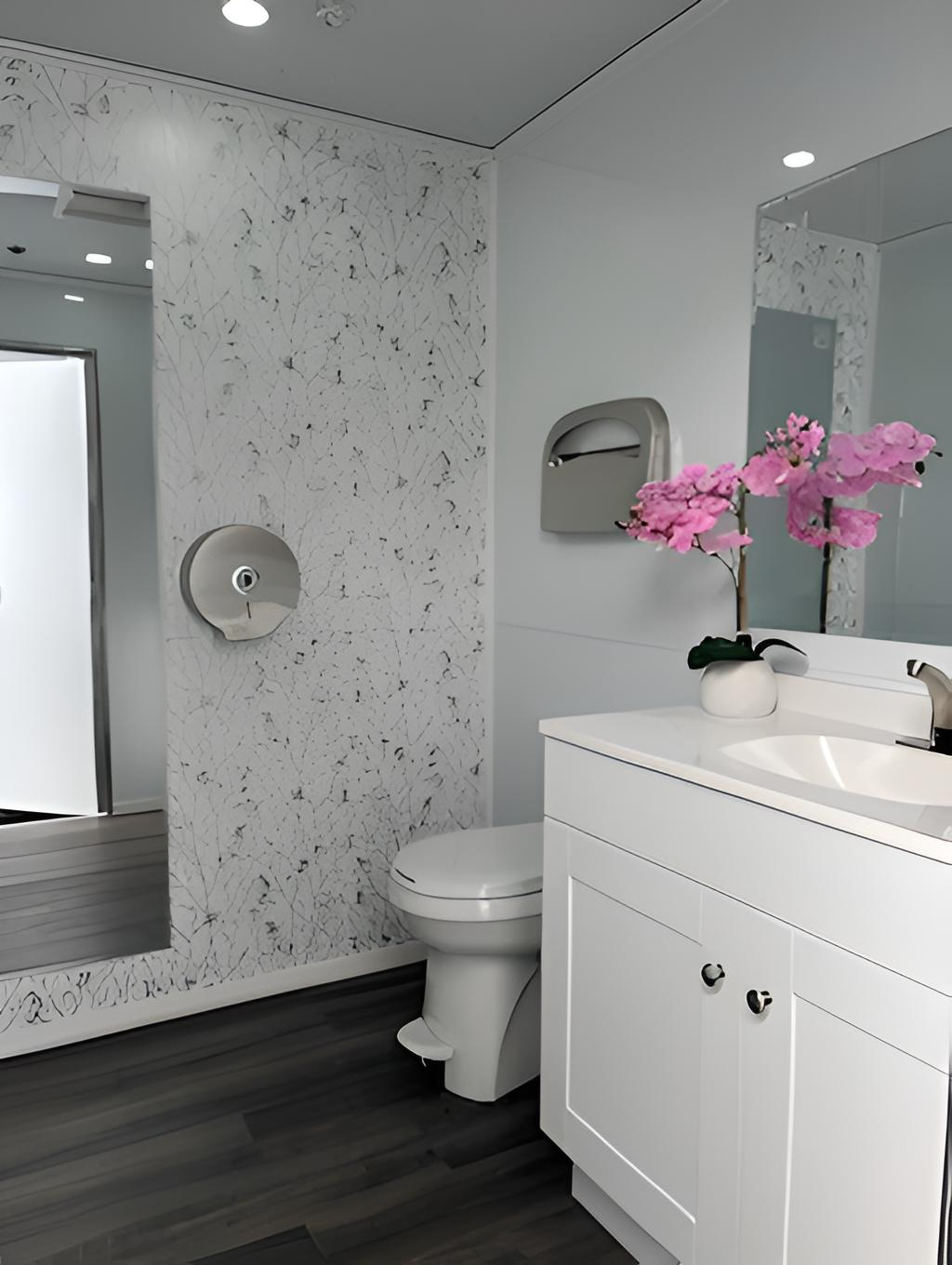 Modern bathroom with white vanity, pink orchid in white vase, and wall-mounted toilet. Features a reflective mirror, a stainless steel toilet paper dispenser, and a granite-patterned divider wall. Grey wood flooring and ceiling lights complete the decor.