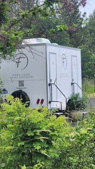 White mobile restroom trailer with Foxy Loops Luxury Restrooms logo, surrounded by green foliage and trees.