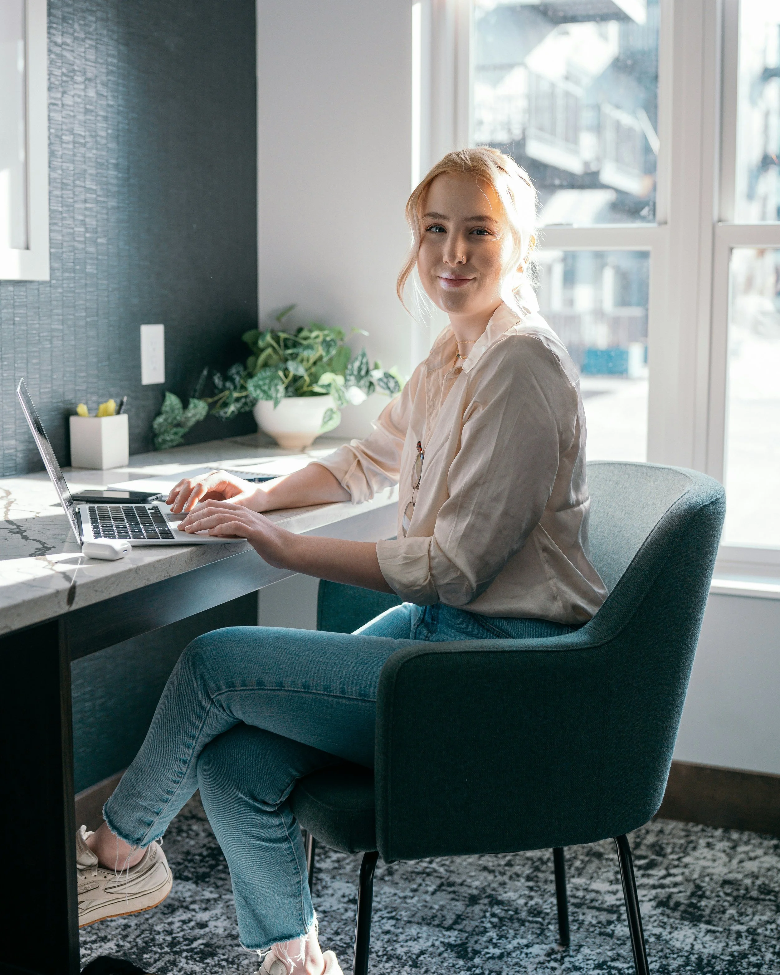 Woman sitting at a desk working on a laptop near a window with sunlight, with plants on the desk.