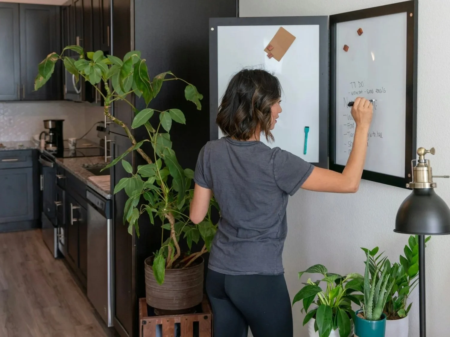 Woman writing on a whiteboard in a kitchen with green plants and black cabinets.