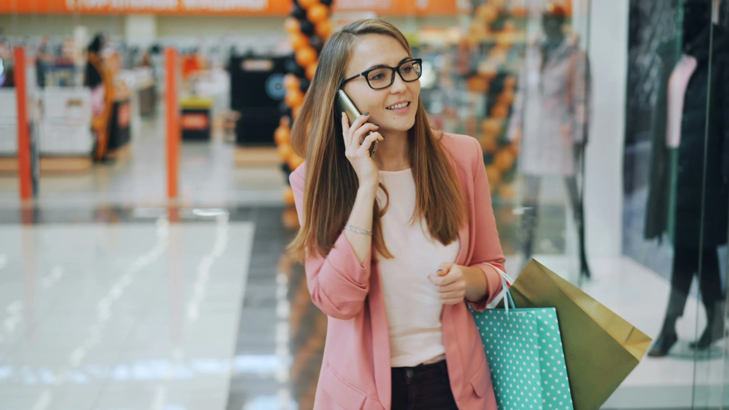 A young woman with long brown hair, wearing glasses, a pink blazer, and a white shirt, is talking on her cellphone while holding shopping bags in a store.