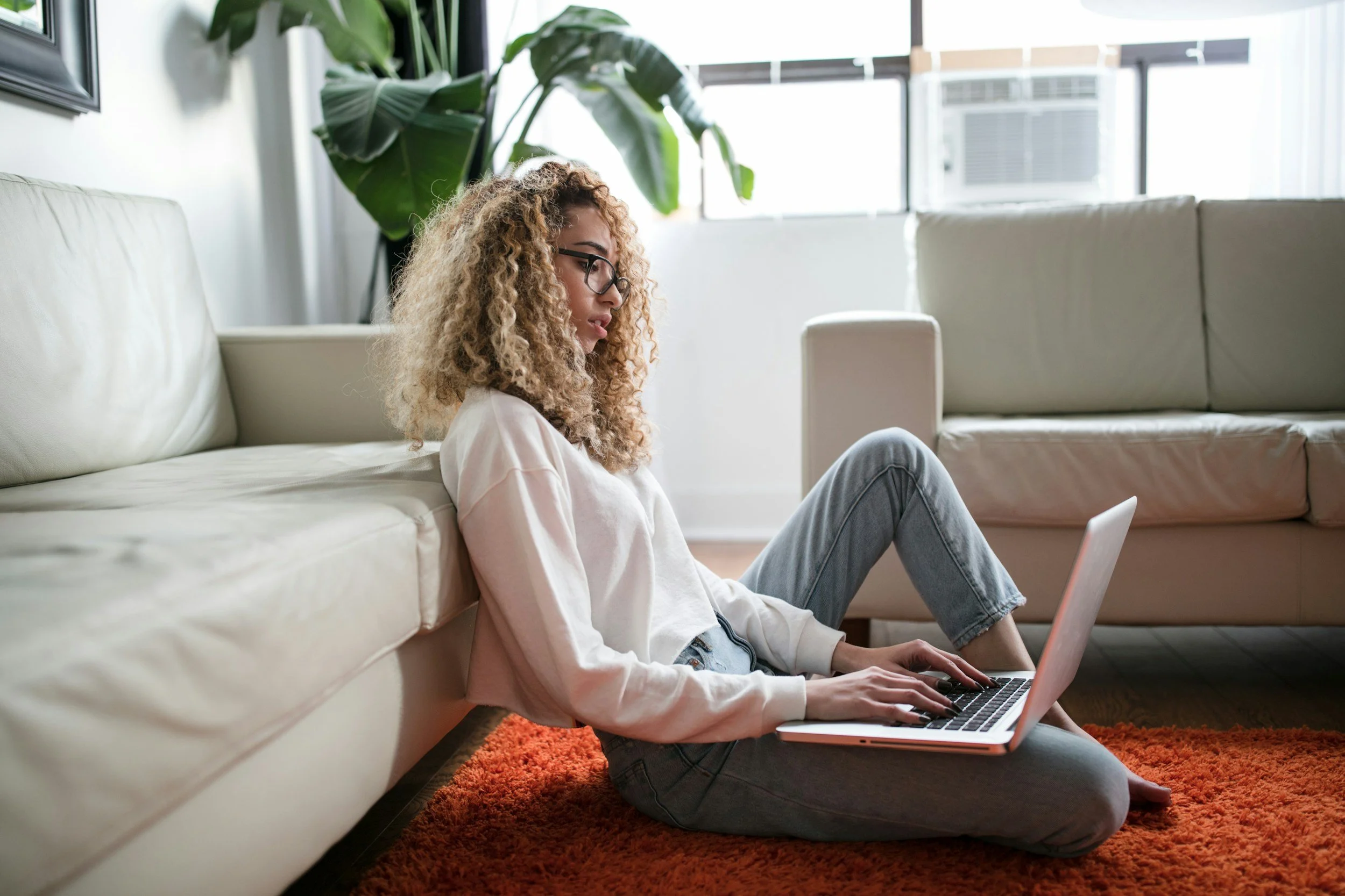 Young woman with curly hair, glasses, wearing a white sweatshirt and jeans, sitting on a orange shag rug, working on a laptop in a bright living room with large windows, beige sofa, and a tall plant.