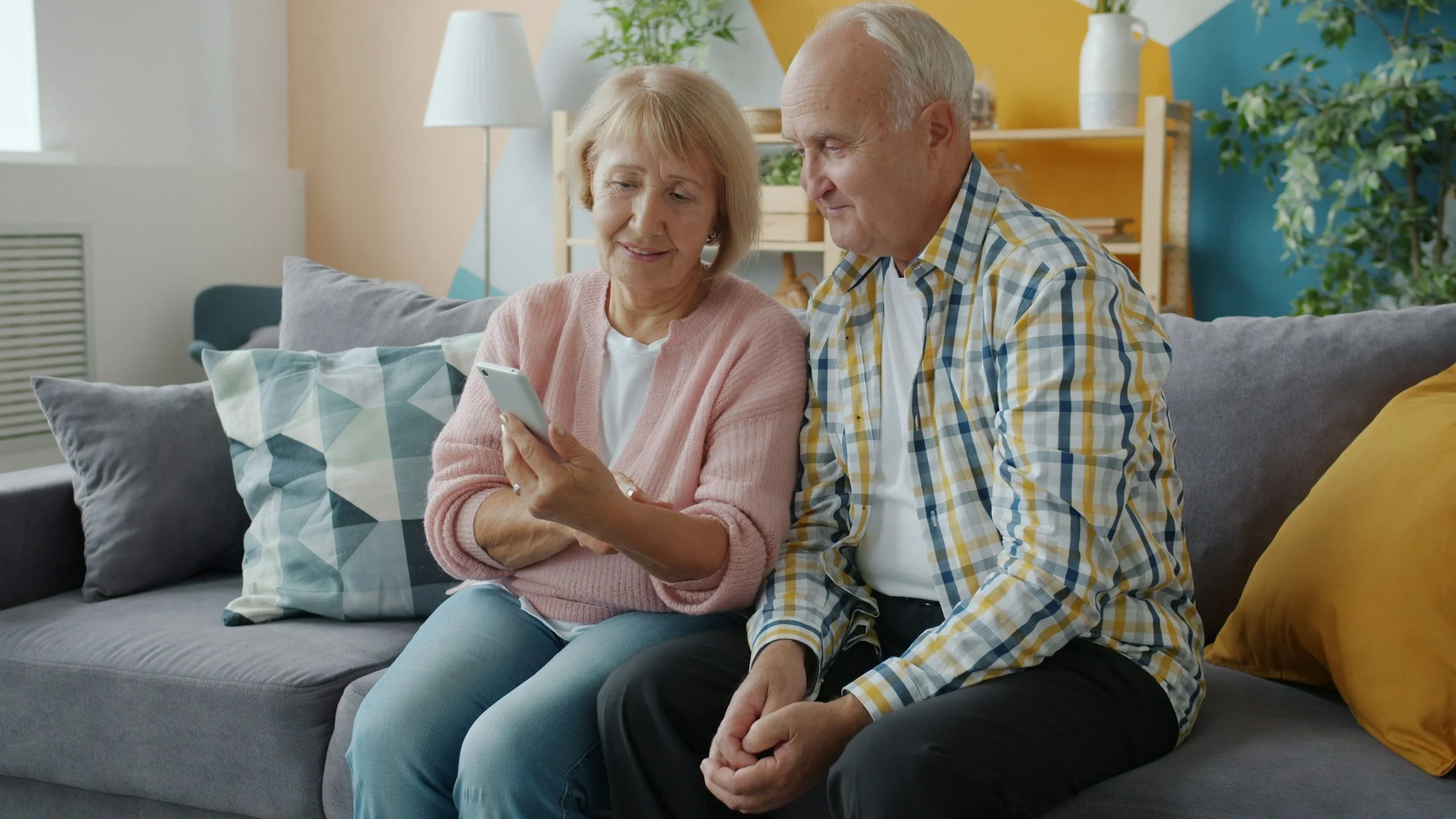 An elderly woman and man sitting on a gray sofa, looking at a smartphone together, in a colorful living room with pillows, plants, and decorative items.