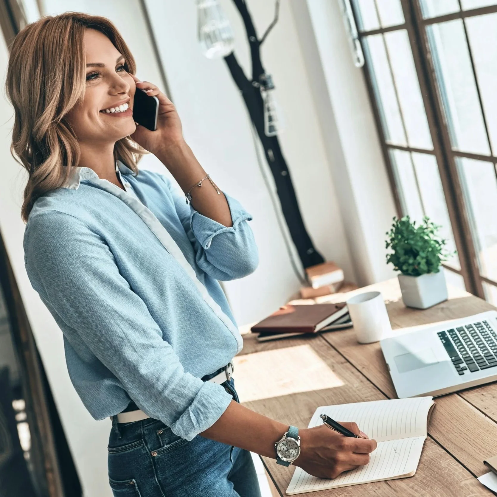 woman writing notes at dining table while on the phone