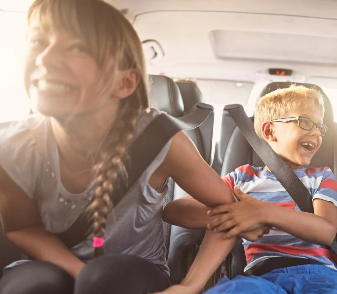 Two children laughing and playing inside a car, with one pulling on the other's arm, both wearing seat belts.