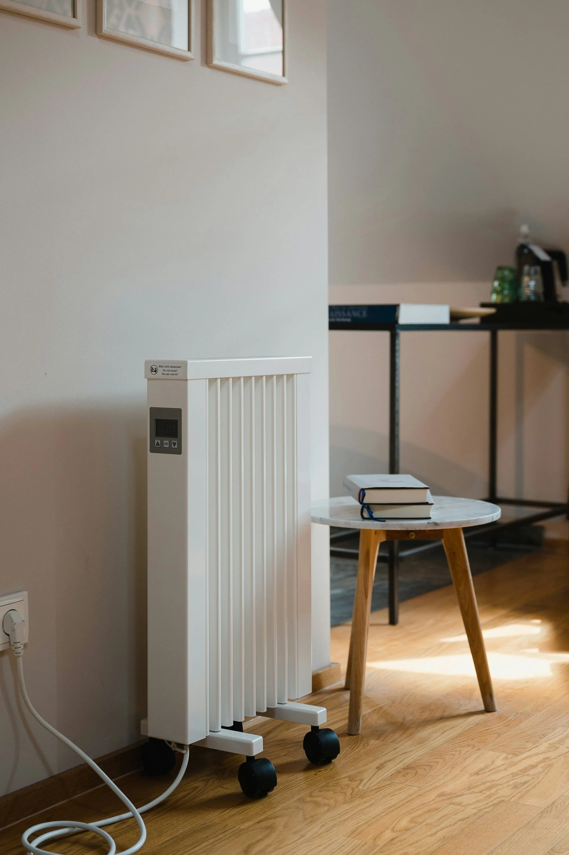 A white portable electric radiator with a digital display, two control buttons, and wheels, placed next to a small wooden round table with three stacked books, in a room with wooden flooring and a black desk in the background.