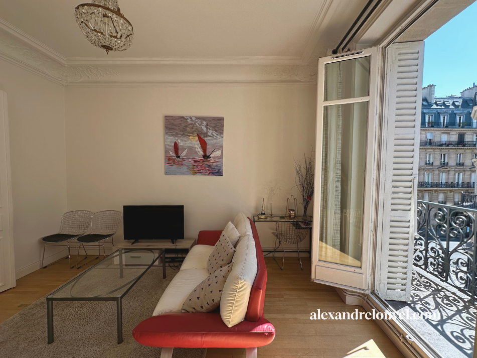 Salon parisien avec canapé, deux chaises, télévision, et balcon donnant sur des immeubles typiques de Paris.