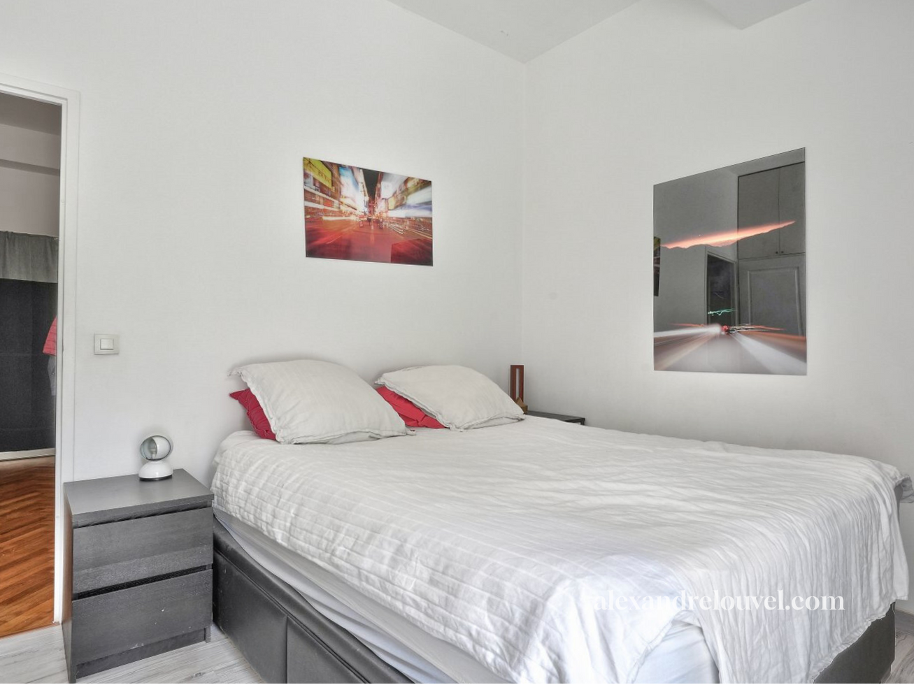 Chambre avec lit double à draps blancs, deux oreillers, deux tableaux modernes au mur, porte ouverte vers un autre pièce, sol en bois.