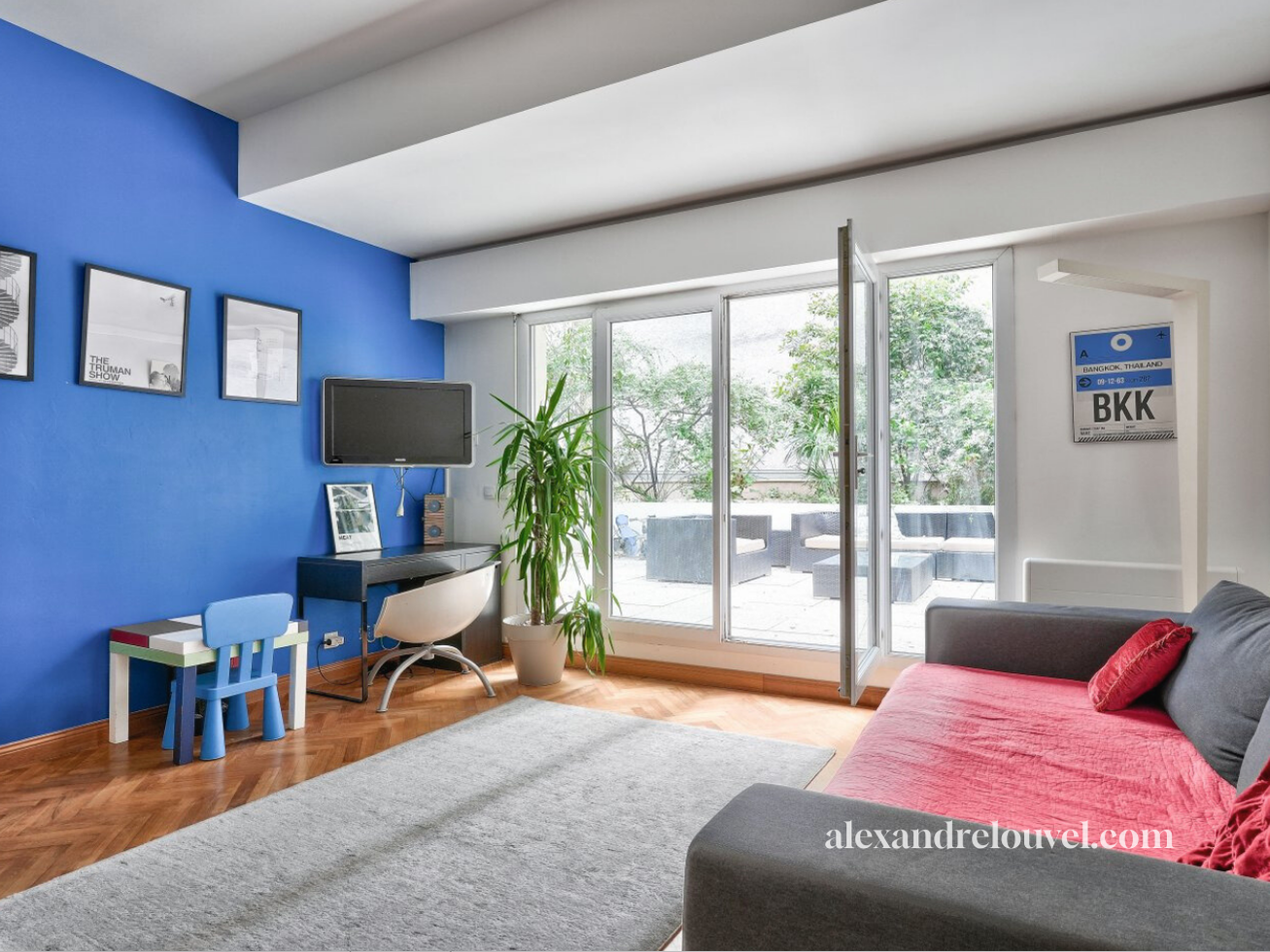 Salon avec canapé gris et rouge, porte-fenêtre donnant sur un balcon avec des meubles d'extérieur, mur blanc avec un tableau d'affichage BKK, télévision, meuble avec enceinte et photo, plante verte, tapis gris, enfants jouant devant la télévision