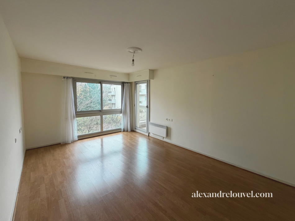 Salon vide avec grandes fenêtres, parquet en bois clair, murs blancs, et une porte-fenêtre menant à un balcon