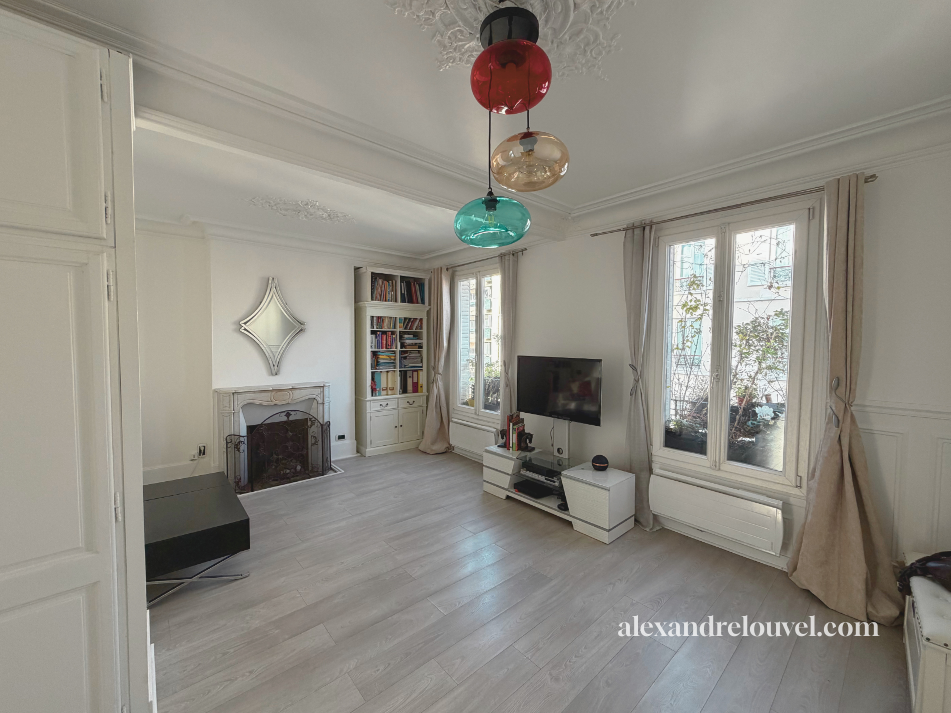 Salon lumineux avec trois fenêtres, un miroir au-dessus d'une cheminée, une télévision sur un meuble blanc, une étagère remplie de livres, et un plafond décoré avec un lustre coloré composé de globes en verre rouge, blanc et bleu.