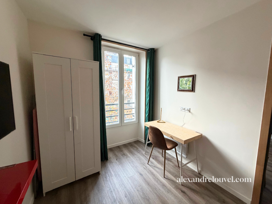 Une chambre avec une armoire blanche, un bureau en bois, une chaise, une fenêtre avec rideaux verts, un tableau accroché au mur, et un sol en bois.