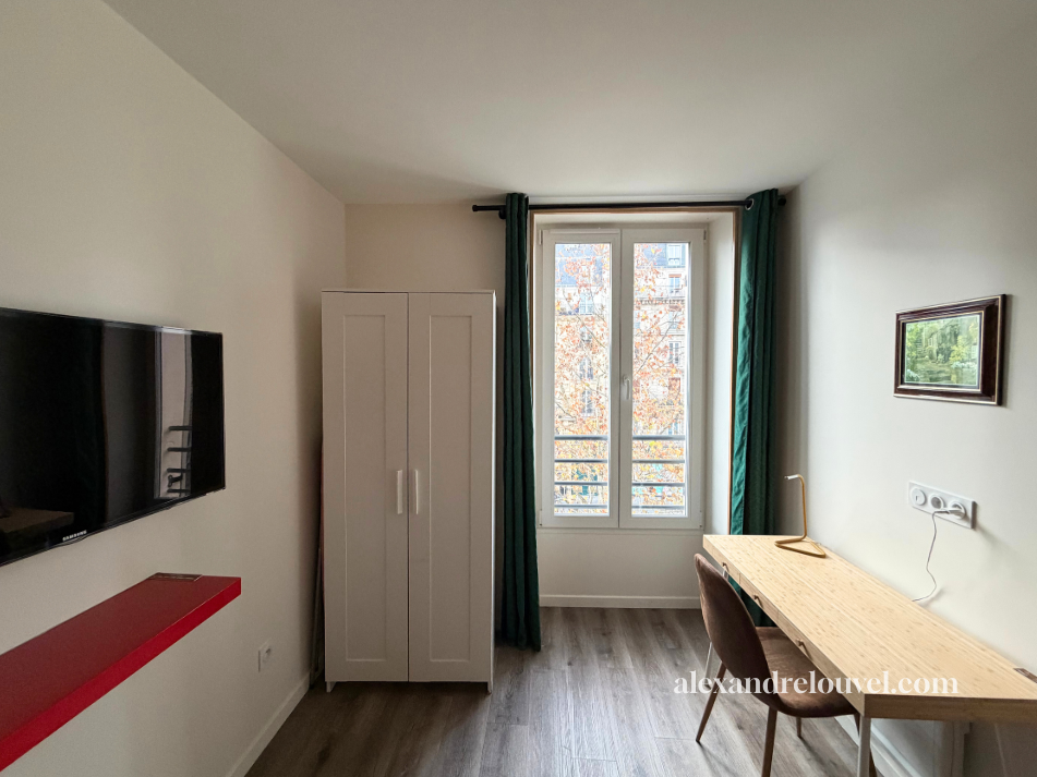 Une pièce d'un appartement avec une télévision, une armoire blanche, une fenêtre avec des rideaux verts, un bureau en bois avec une lampe, et une chaise. La pièce est lumineuse avec vue sur des arbres.