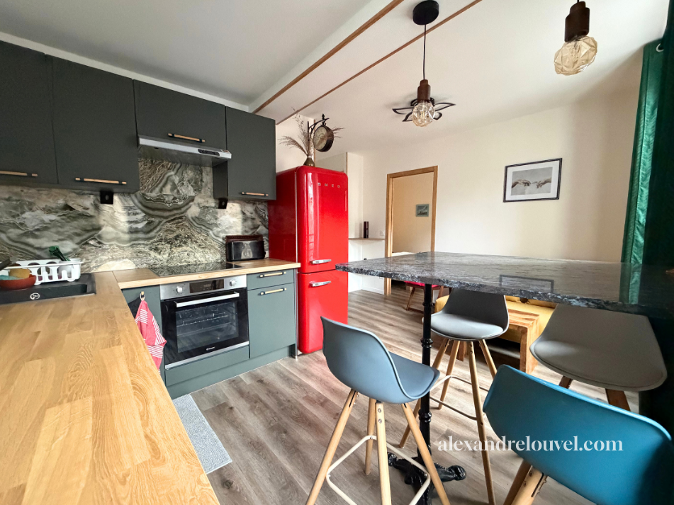 Cuisine moderne avec plan de travail en granit, armoires vertes, réfrigérateur rouge, rangements, table en bois et chaises modernes, décoration murale et éclairage suspendu.