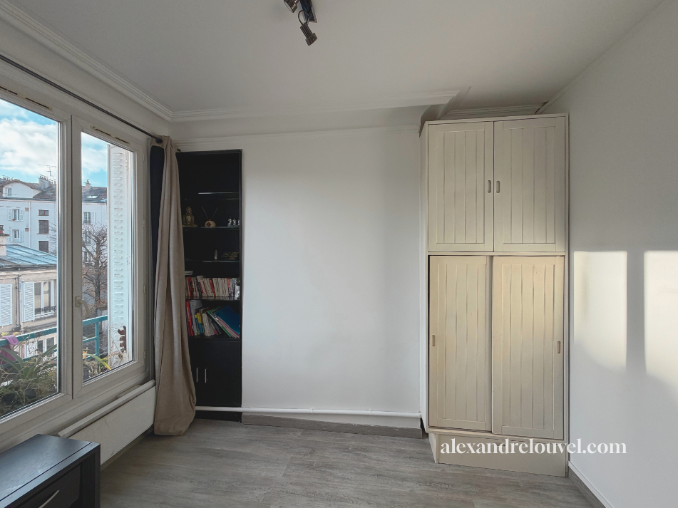 Une pièce lumineuse avec un grand fenêtre, une armoire en bois clair, une étagère noire avec des livres et des décorations, et un sol en bois clair.