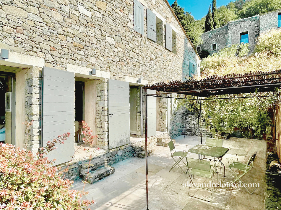 Cour intérieure avec une maison en pierre, fenêtres avec volets gris, un mobilier de jardin comprenant une table et des chaises, un pergola en bois, et des plantes vertes en arrière-plan.