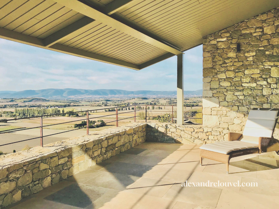 Balcon avec une chaise longue en rotin, vue sur un paysage rural avec champs, arbres et montagnes à l'horizon, sous un ciel partiellement nuageux.
