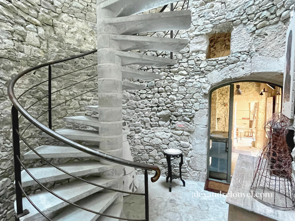 Escalier en spirale en béton dans une pièce avec mur en pierre naturelle, porte en verre, et mobilier vintage.