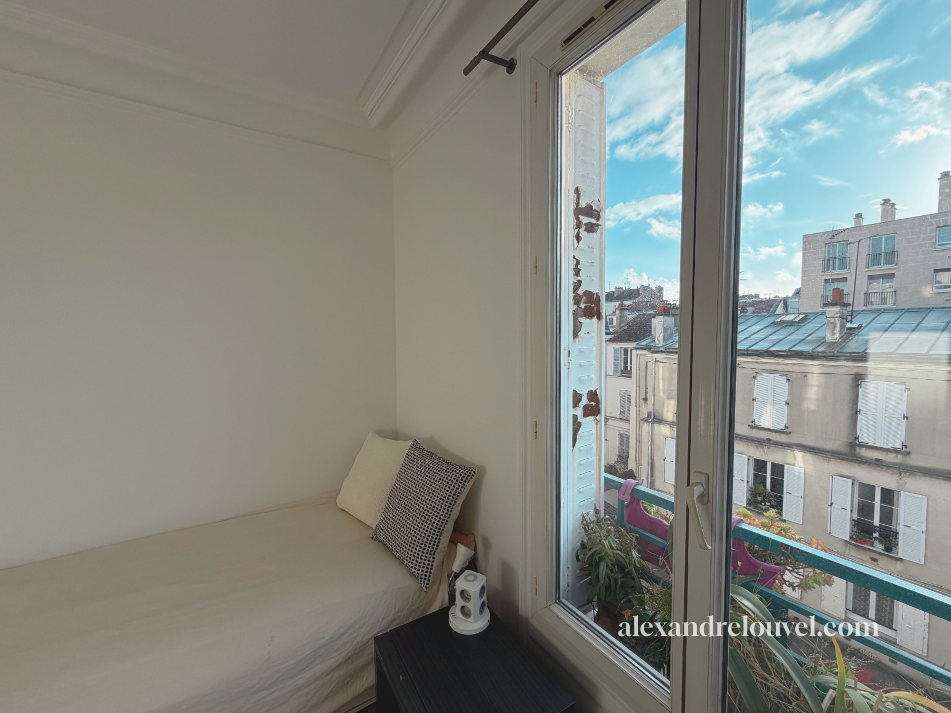 Chambre avec un lit simple, deux coussins, un petit table de chevet avec une enceinte, un mur blanc, une grande fenêtre donnant sur des bâtiments parisiens, un ciel bleu ensoleillé.