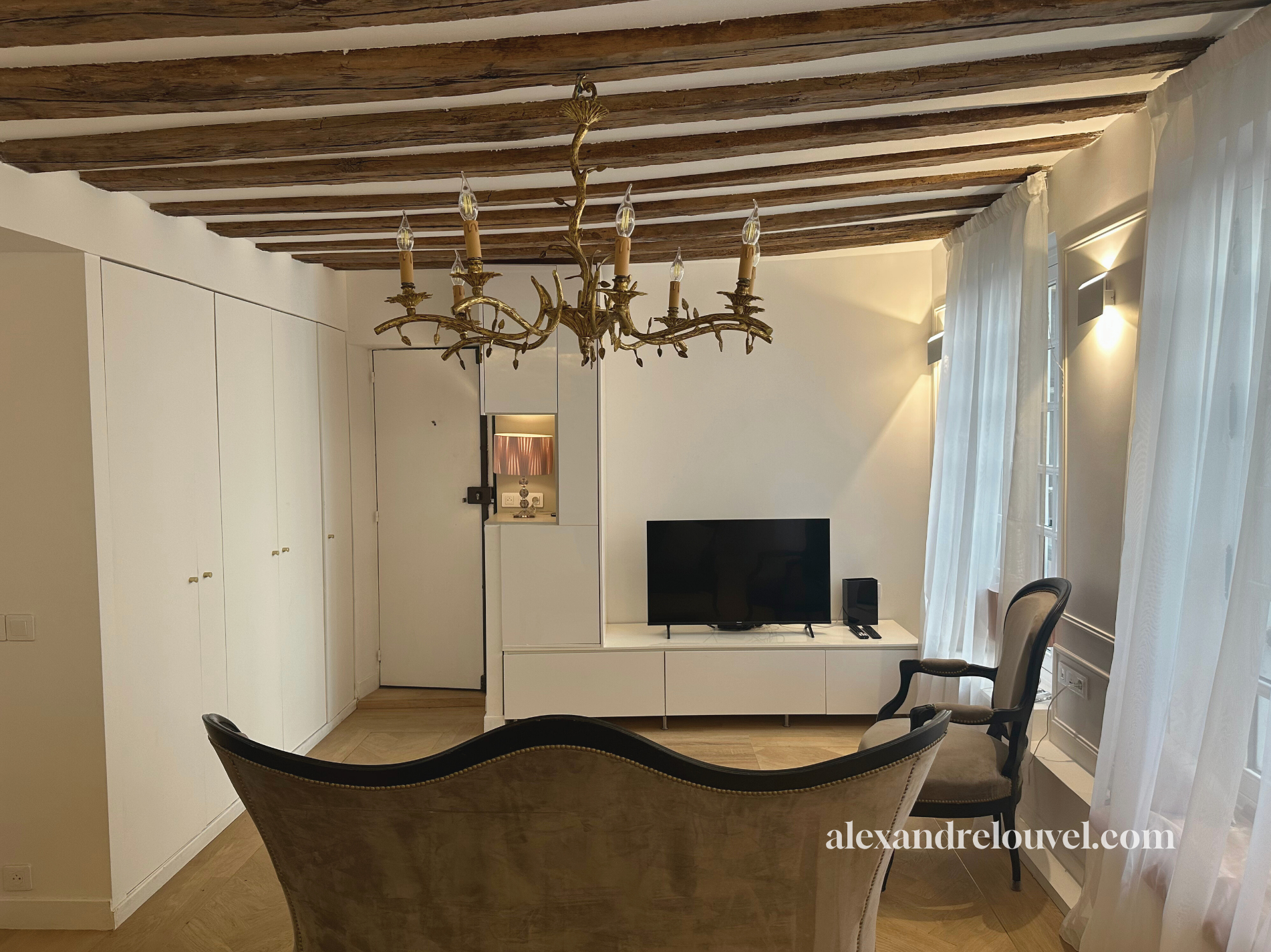 Salon élégant avec un plafond en bois apparent, un chandelier en métal doré, une télévision sur un meuble blanc, deux chaises anciennes, rideaux blancs et murs beige clair.