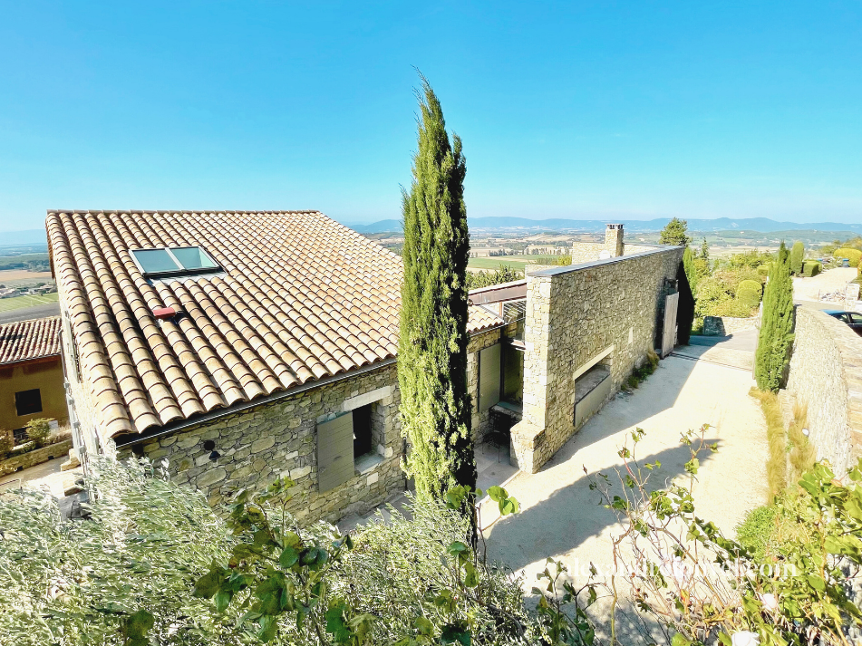 Une maison en pierre avec un toit en tuiles, entourée de sapins, dans un paysage rural ensoleillé.